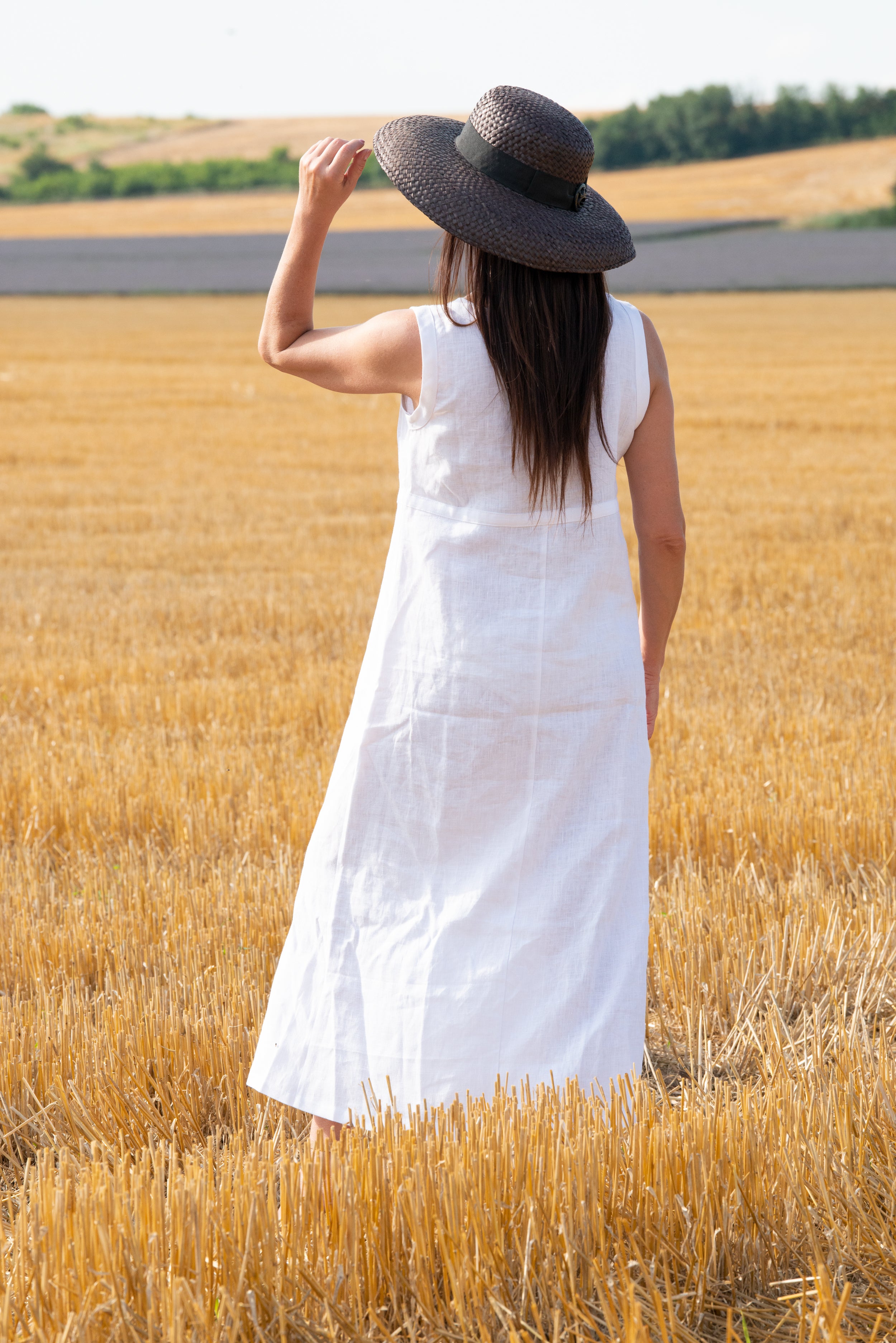 Woman in a white linen dress and straw hat standing in a wheat field.