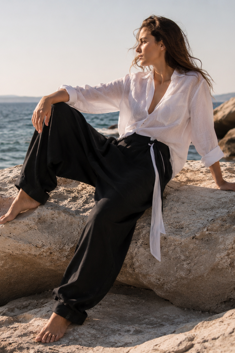 Woman sitting on a rock by the ocean wearing a white shirt and black harem linen pants.