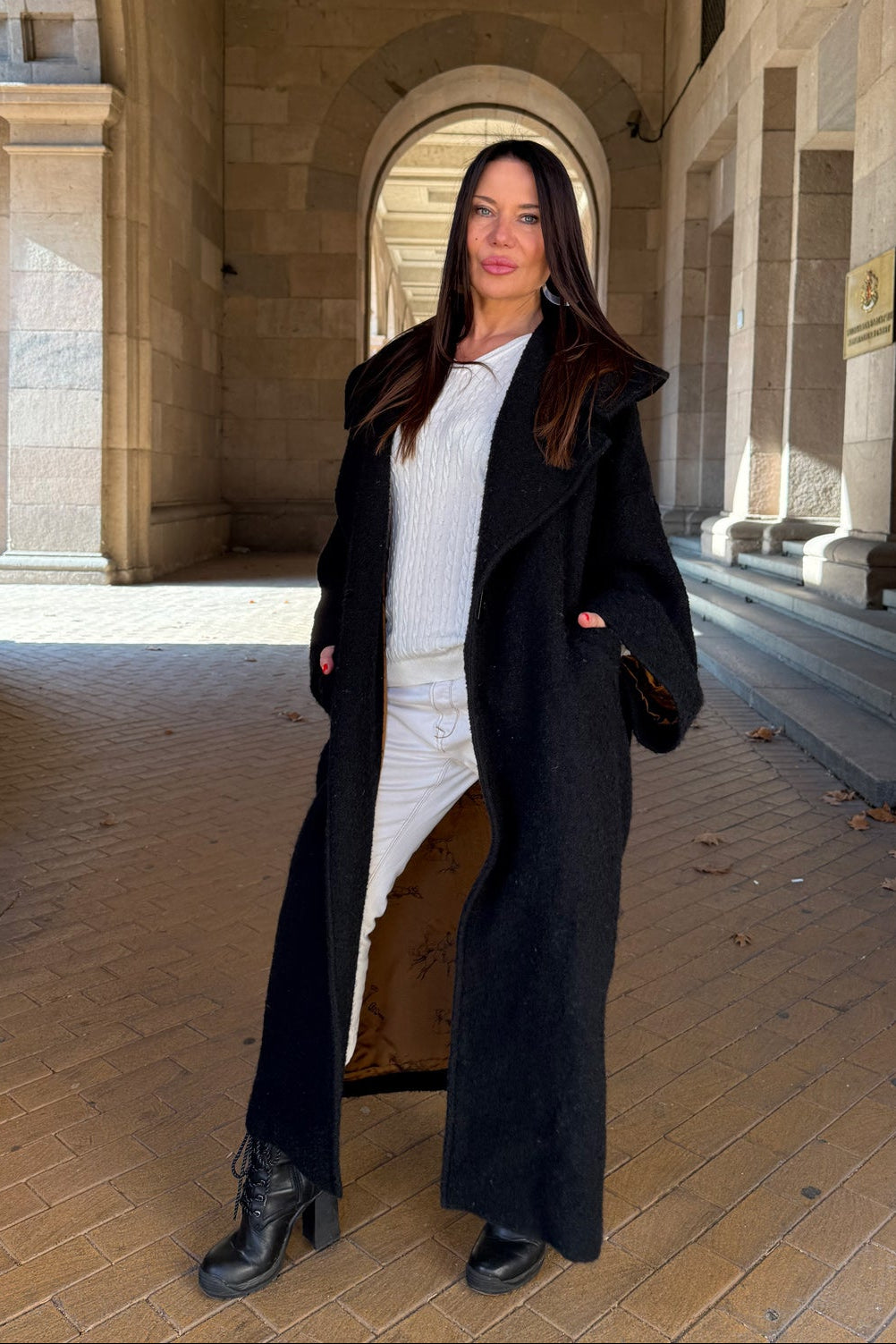 Woman in a long black coat standing in a sunlit stone hallway