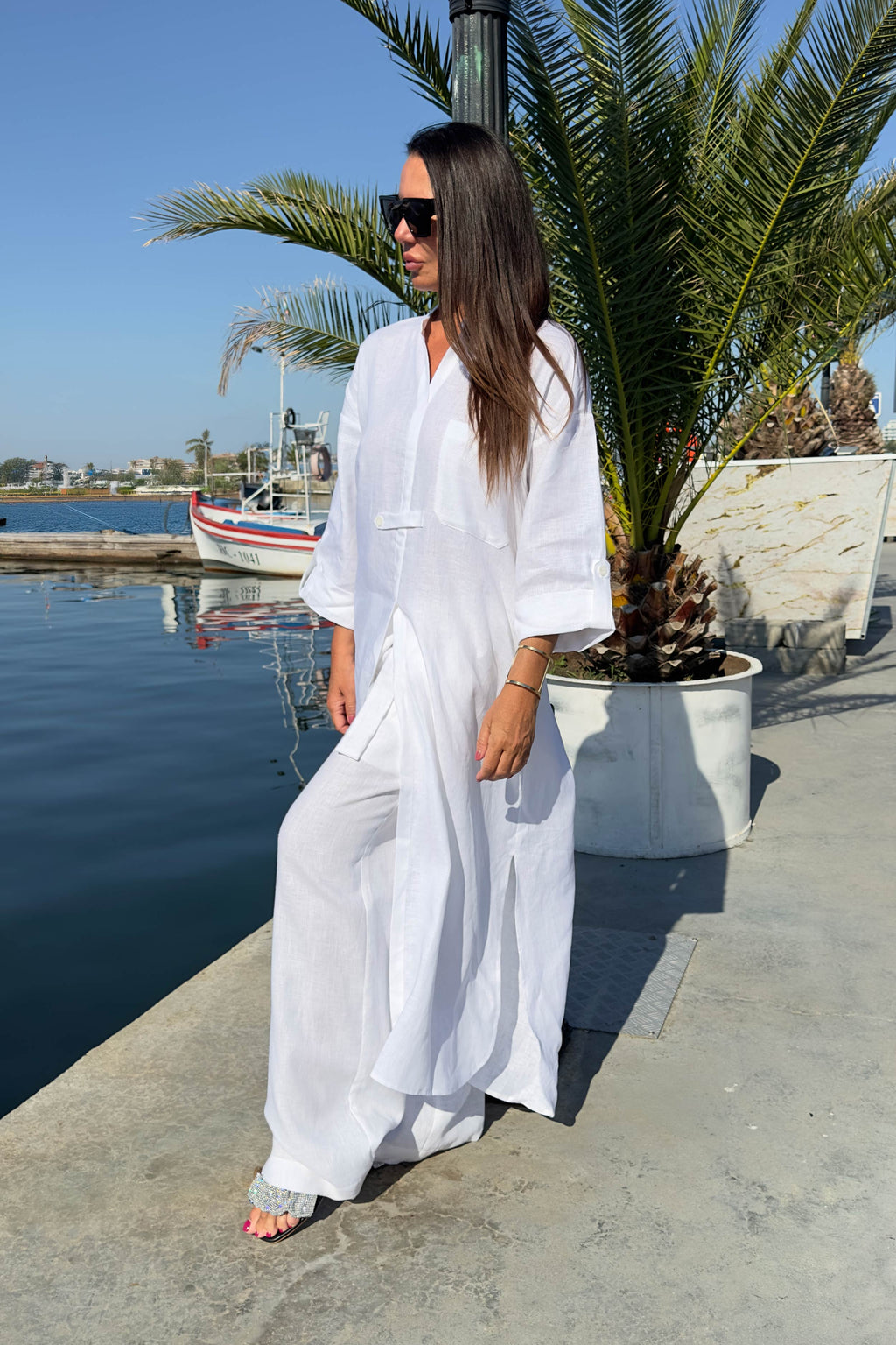 Woman in a white outfit standing by a waterfront with palm trees and a boat in the background.