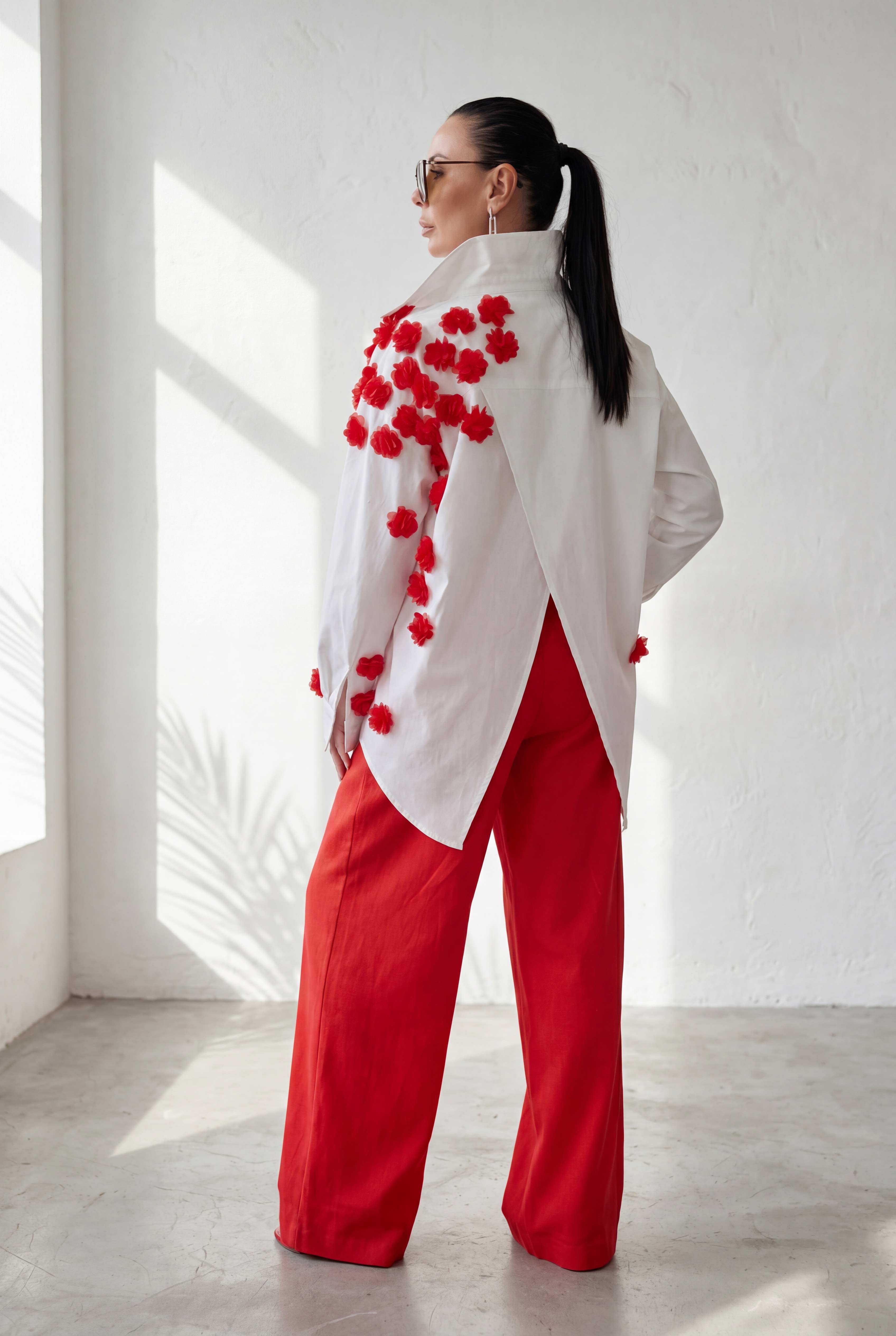 Woman wearing a white top with red floral patterns and red pants in a bright room.