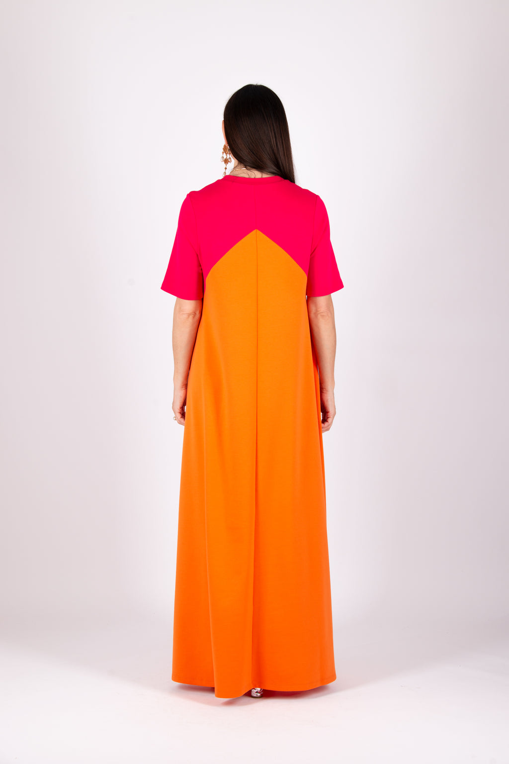 A woman viewed from the back wearing a color-block maxi dress with a bright pink upper yoke and a large orange skirt, photographed against a plain light gray studio background.