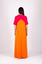 A woman viewed from the back wearing a color-block maxi dress with a bright pink upper yoke and a large orange skirt, photographed against a plain light gray studio background.