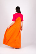 A woman viewed from behind wearing a long color-block dress with a bright pink upper section and a flowing orange lower section, photographed against a plain light gray studio background.