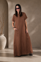 Woman wearing a brown dress standing against a neutral wall with a vase on the left.
