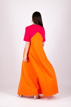 A woman viewed from behind wearing a long color-block dress with a bright orange body and a vivid pink upper yoke and short sleeves, photographed against a plain light gray studio background.