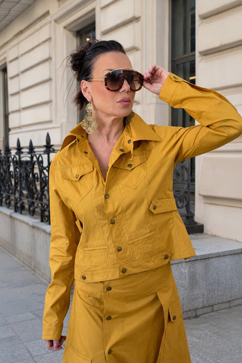Woman in a mustard yellow jacket standing on a street with a building in the background
