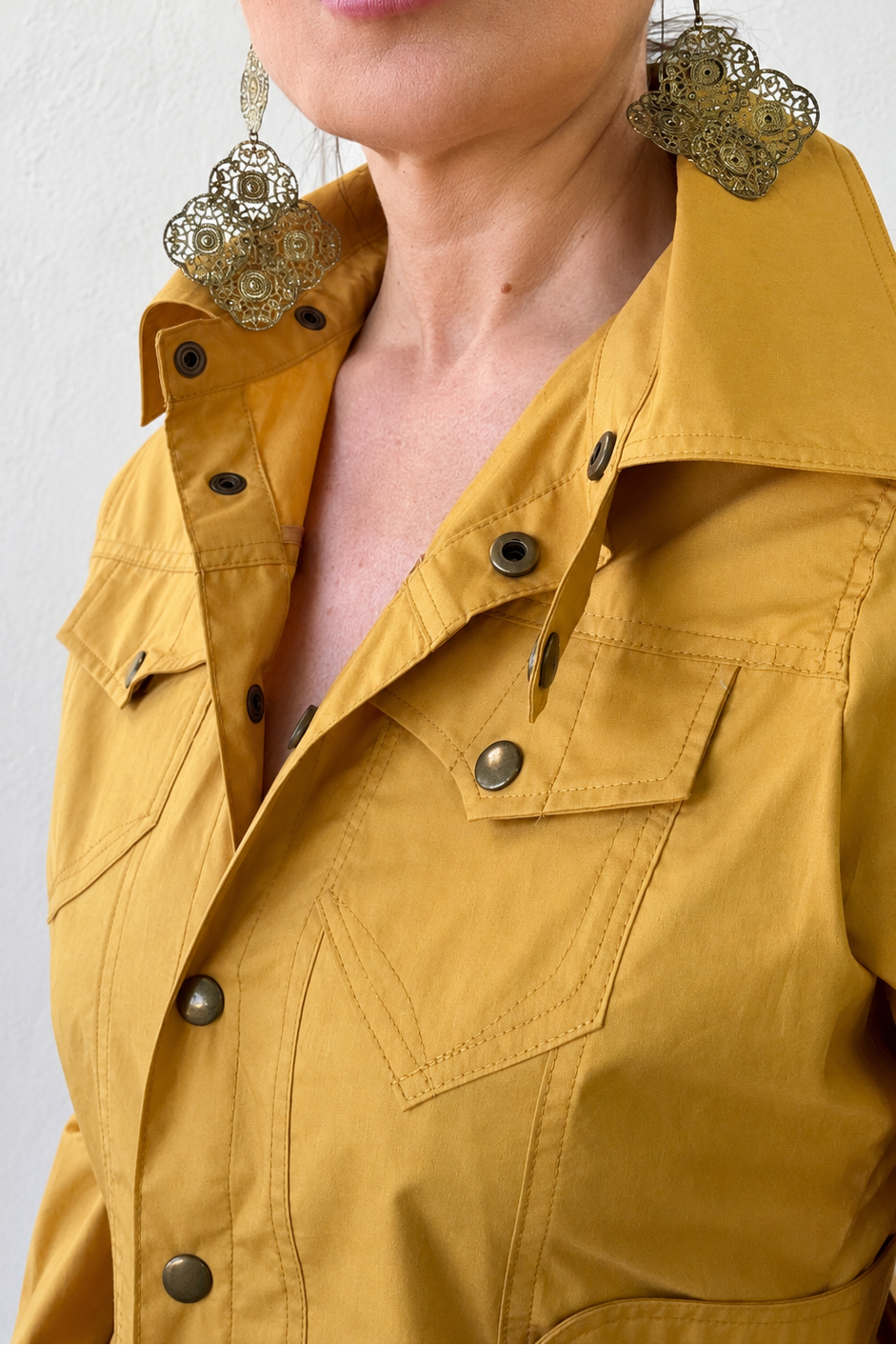 Woman wearing a mustard yellow jacket with large earrings against a white background