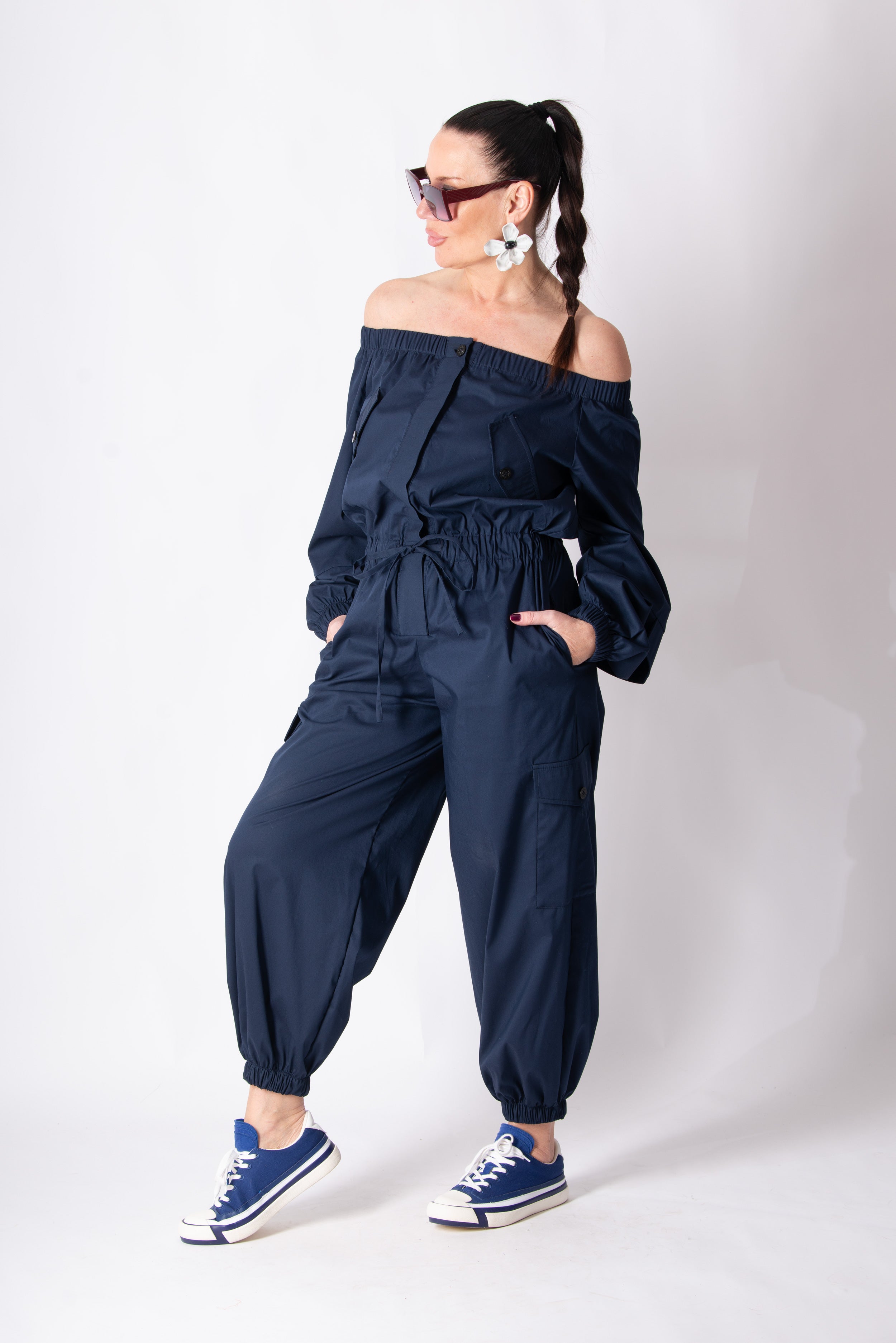 Woman wearing a navy off-shoulder jumpsuit with matching sneakers on a white background