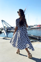 Woman in a polka dot dress standing by a marina with boats and cranes in the background.