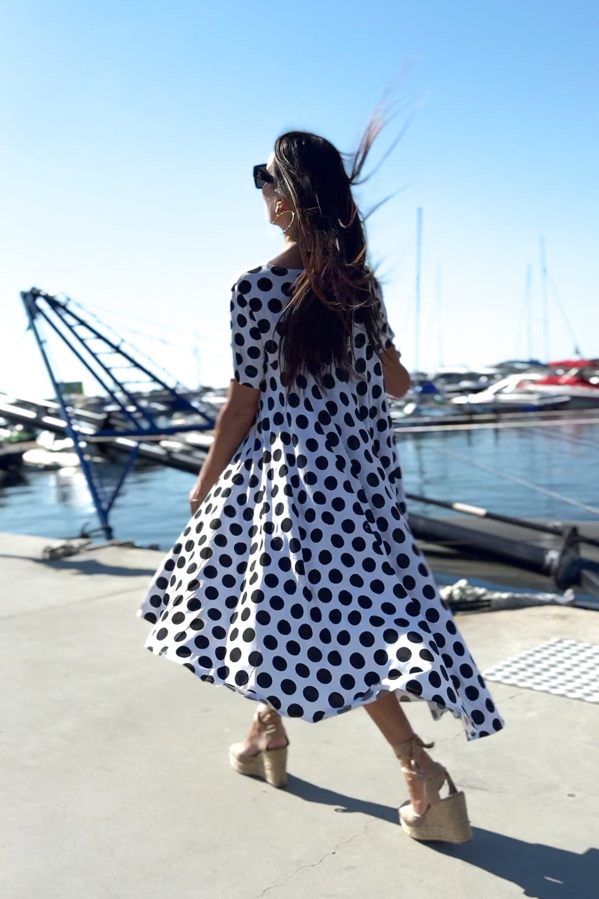 Woman in a polka dot dress standing by a marina with boats and cranes in the background.