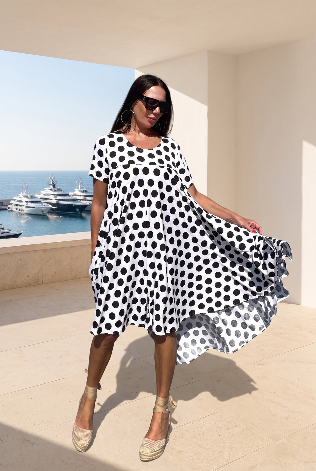 A woman modeling a short-sleeve, knee-length dress with a bold black and white polka dot pattern, standing on a sunlit terrace with a marina and yachts in the background.
