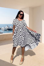 A woman modeling a short-sleeve, knee-length dress with a bold black and white polka dot pattern, standing on a sunlit terrace with a marina and yachts in the background.