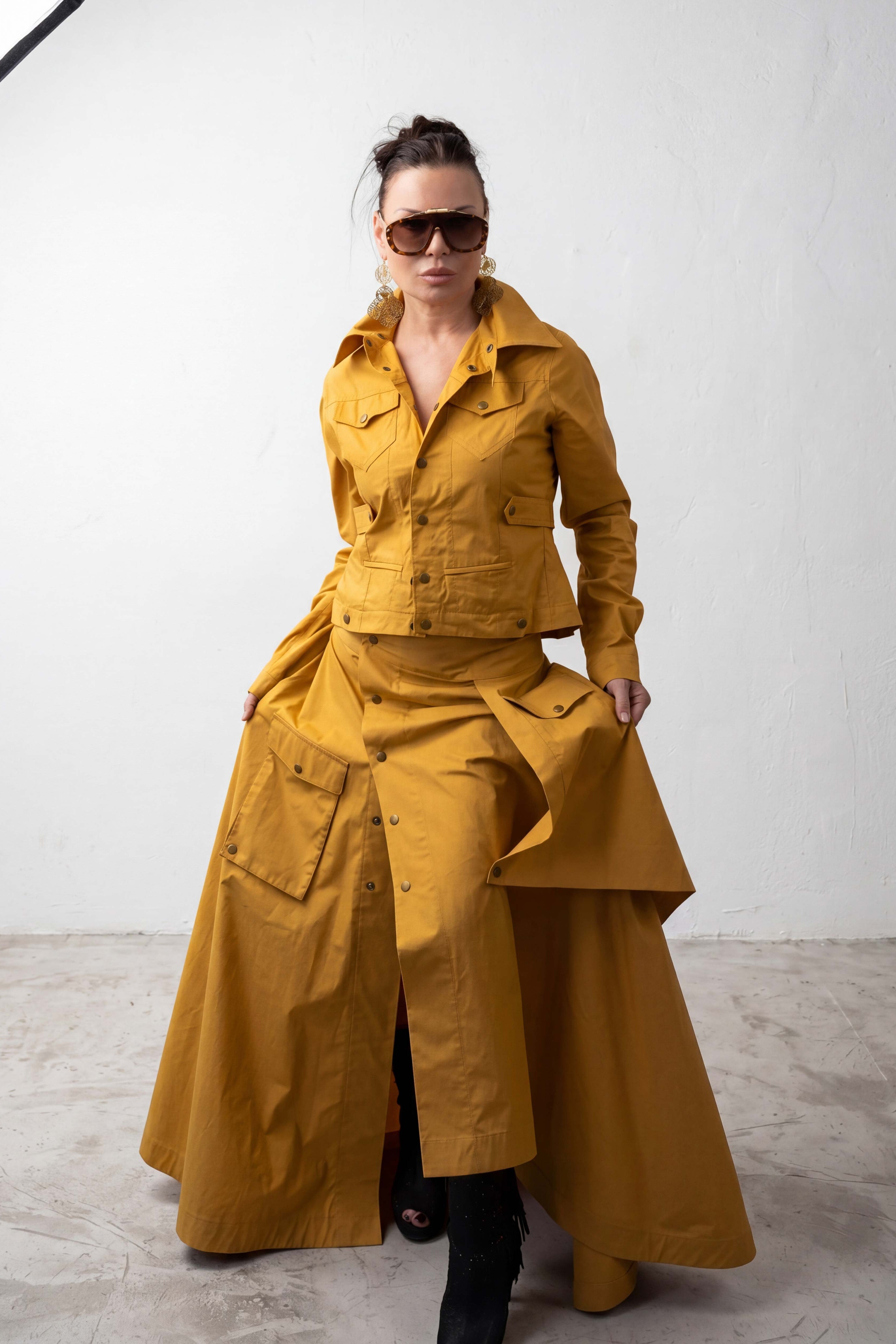 Person wearing a mustard yellow outfit with a long coat and jacket against a white background