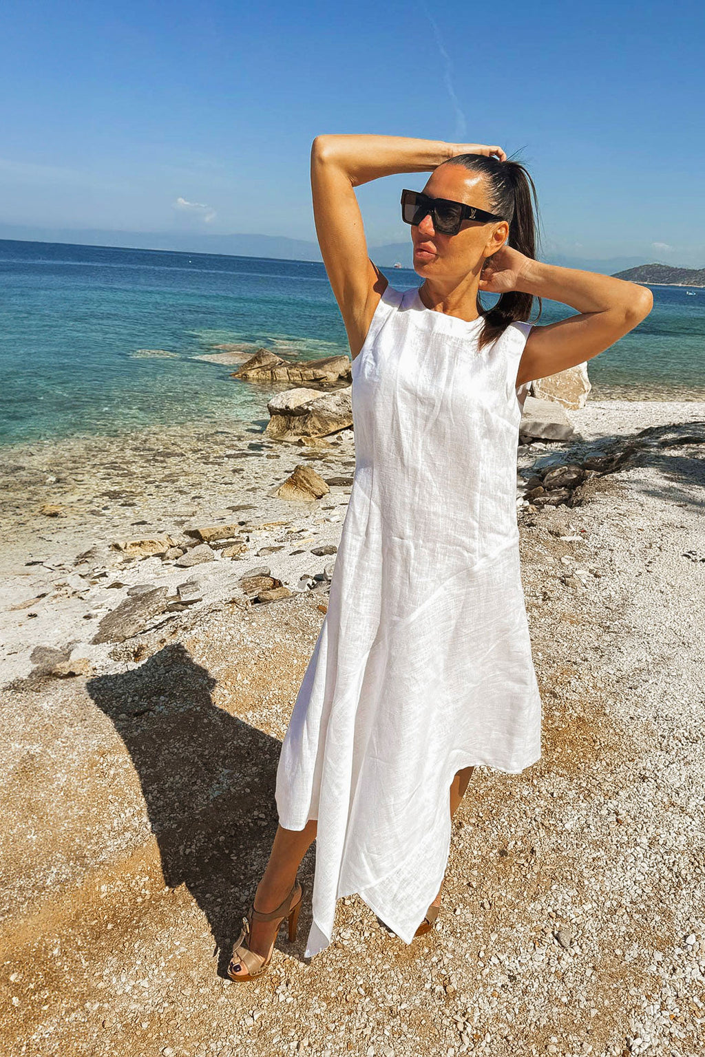 Woman in a white dress standing on a beach with clear blue water and sky.