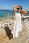Woman in a white dress standing on a beach with clear blue water and sky.