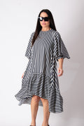 A woman modeling a black and white vertically striped midi dress with a ruffled hem and elbow-length sleeves, styled with black sunglasses against a plain white studio background.