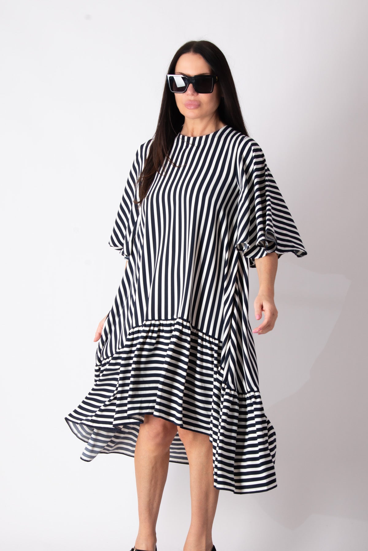 A woman modeling a black and white vertically striped midi dress with a ruffled hem and elbow-length sleeves, styled with black sunglasses against a plain white studio background.