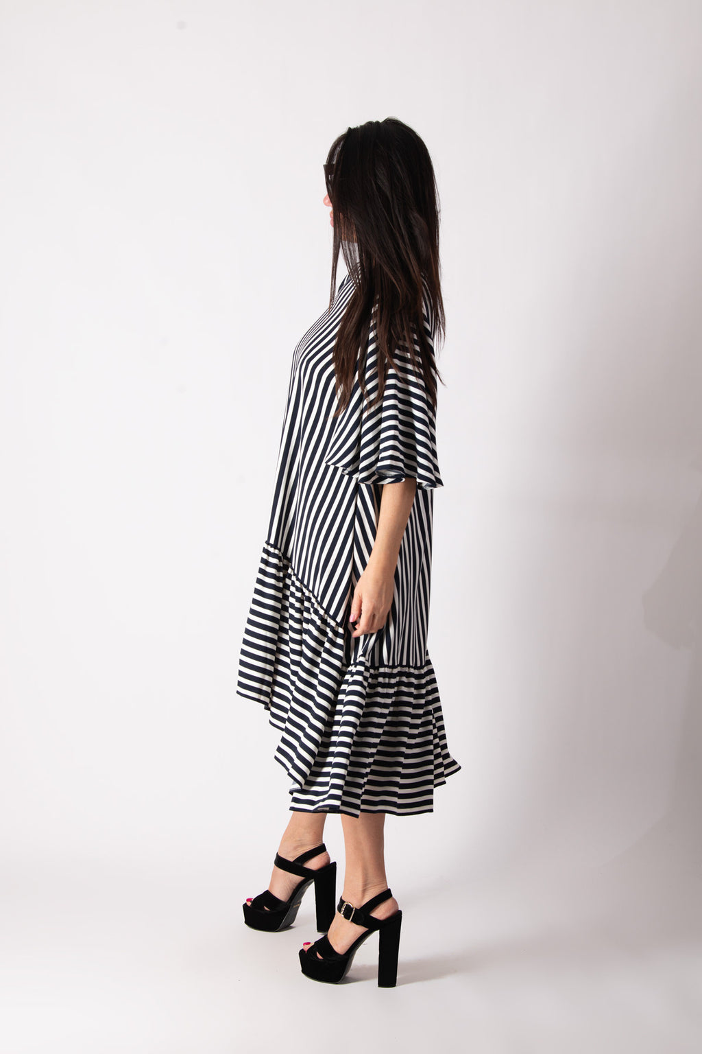 A woman wearing a black and white striped midi dress with tiered ruffle hems and elbow-length sleeves, paired with black platform sandals, shown from a side-back angle against a white studio background.