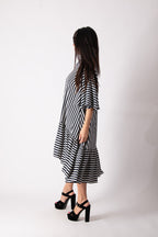 A woman wearing a black and white striped midi dress with tiered ruffle hems and elbow-length sleeves, paired with black platform sandals, shown from a side-back angle against a white studio background.