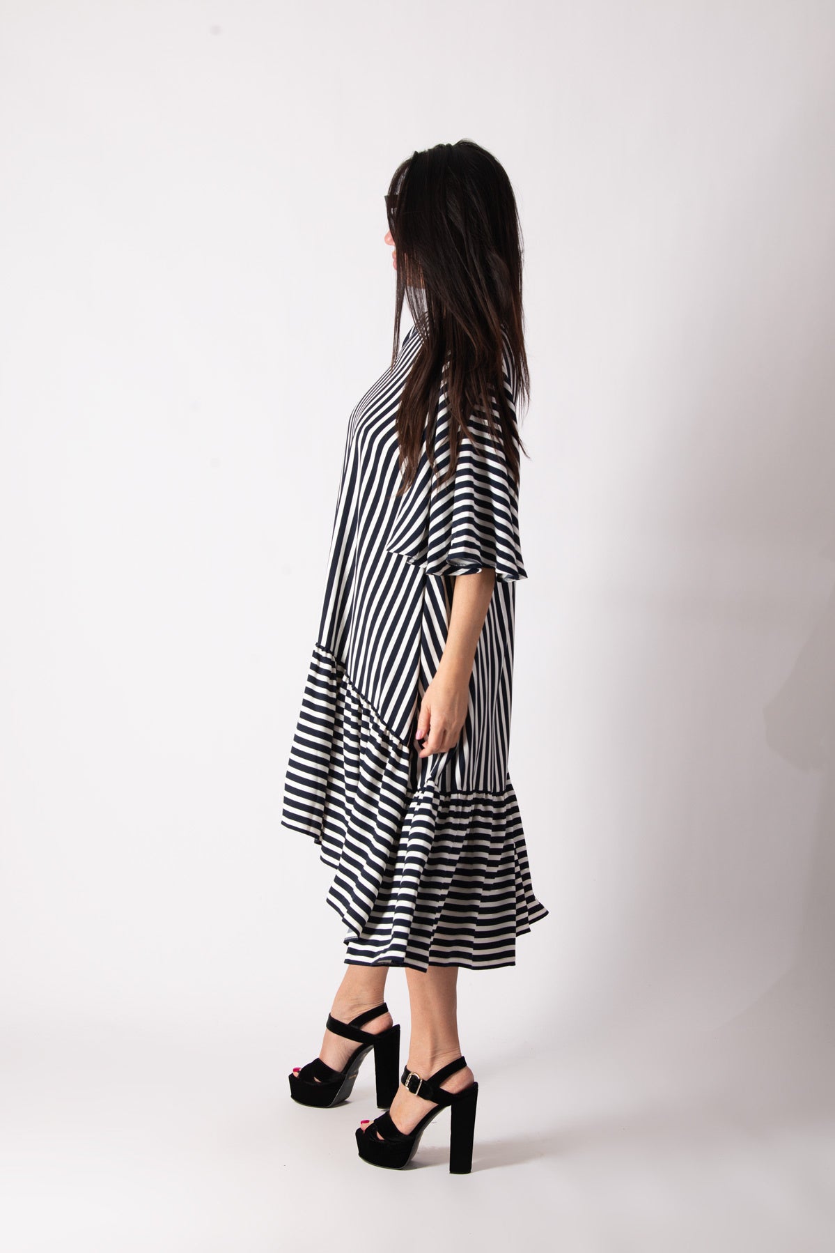 A woman wearing a black and white striped midi dress with tiered ruffle hems and elbow-length sleeves, paired with black platform sandals, shown from a side-back angle against a white studio background.