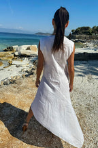 Woman in a white dress walking on a rocky beach with clear blue water and sky.