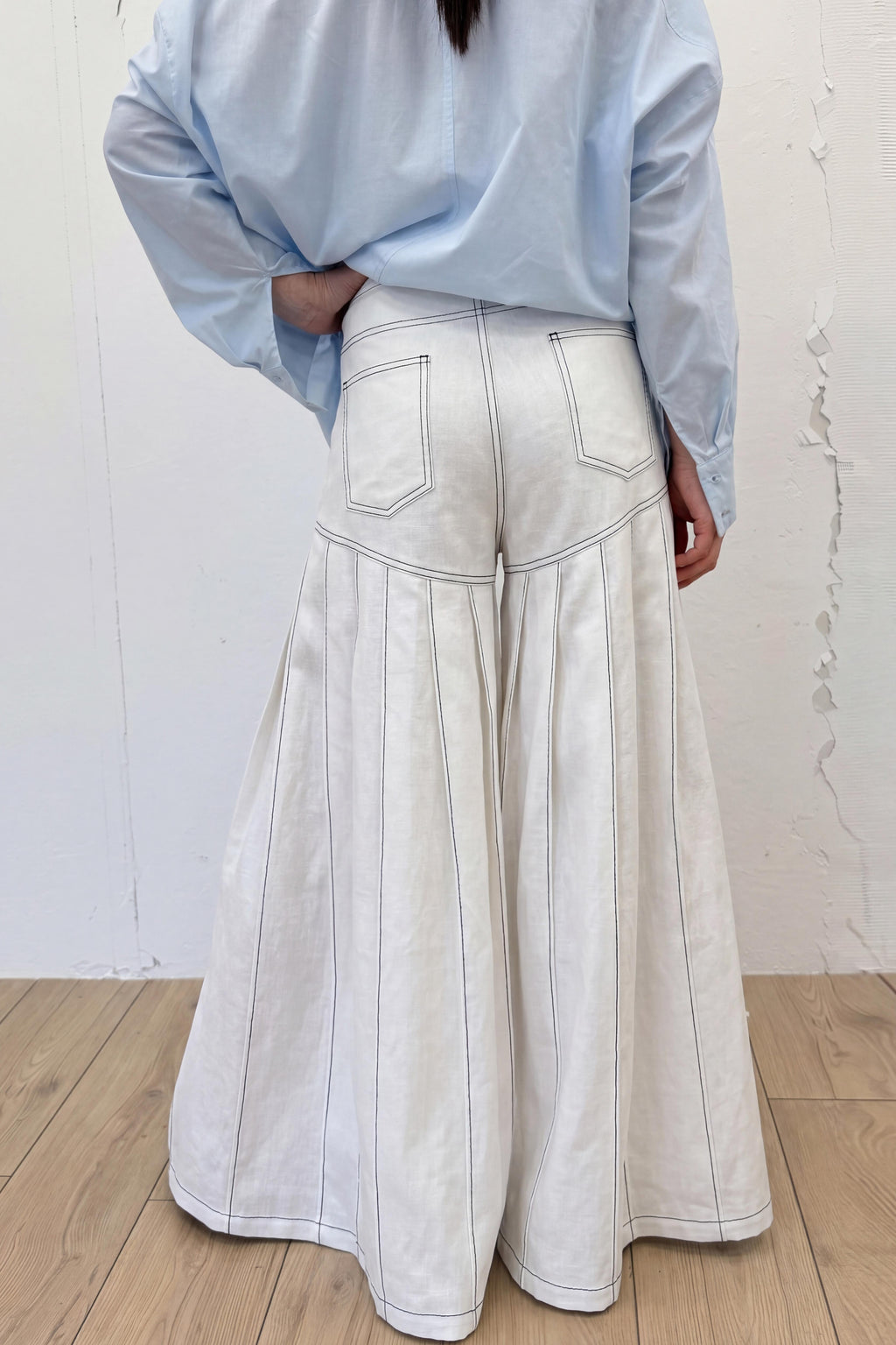 Person wearing a light blue shirt and white wide-leg pants against a white wall.