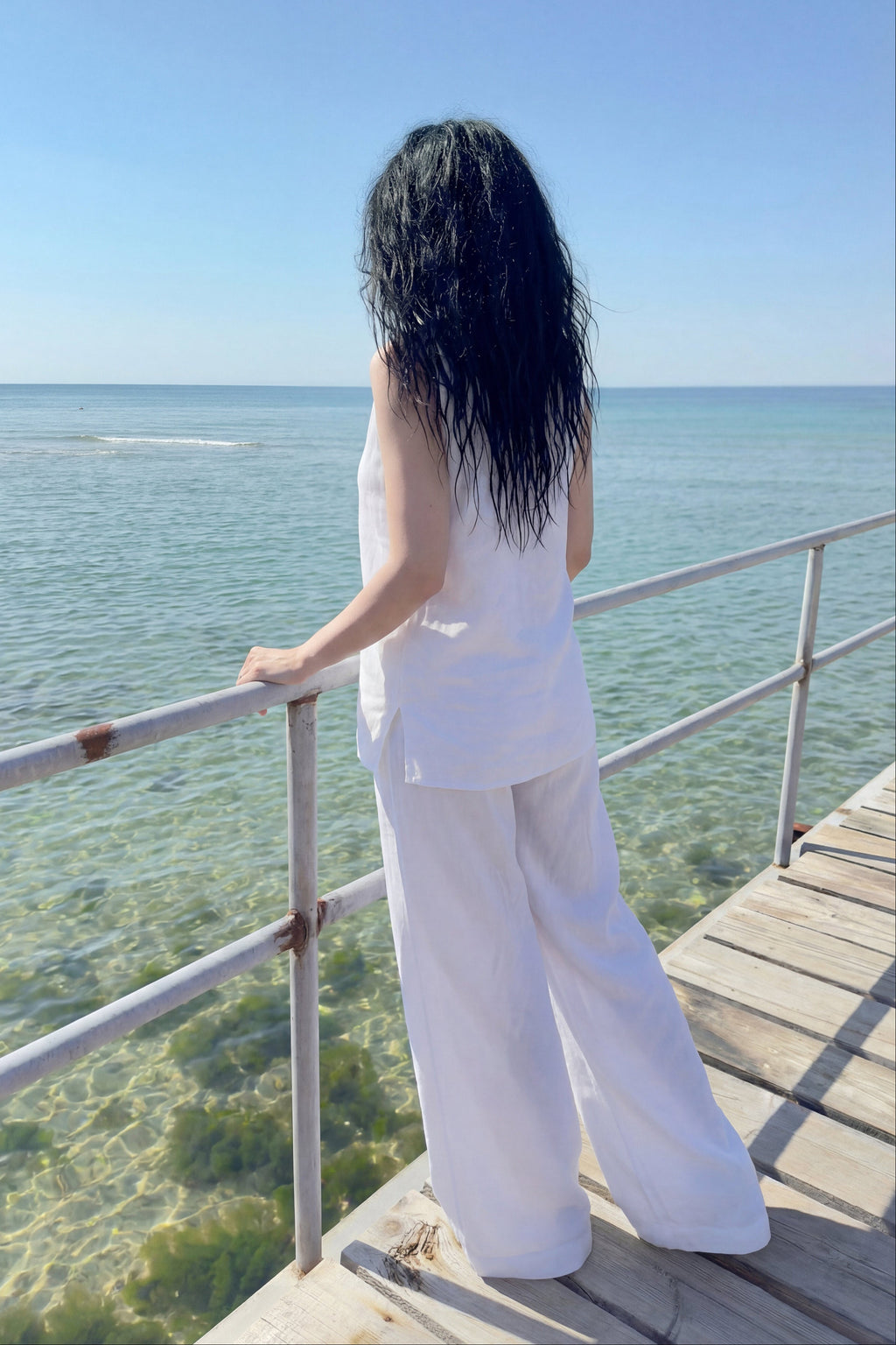 Person in white outfit standing on a wooden deck overlooking clear blue water.