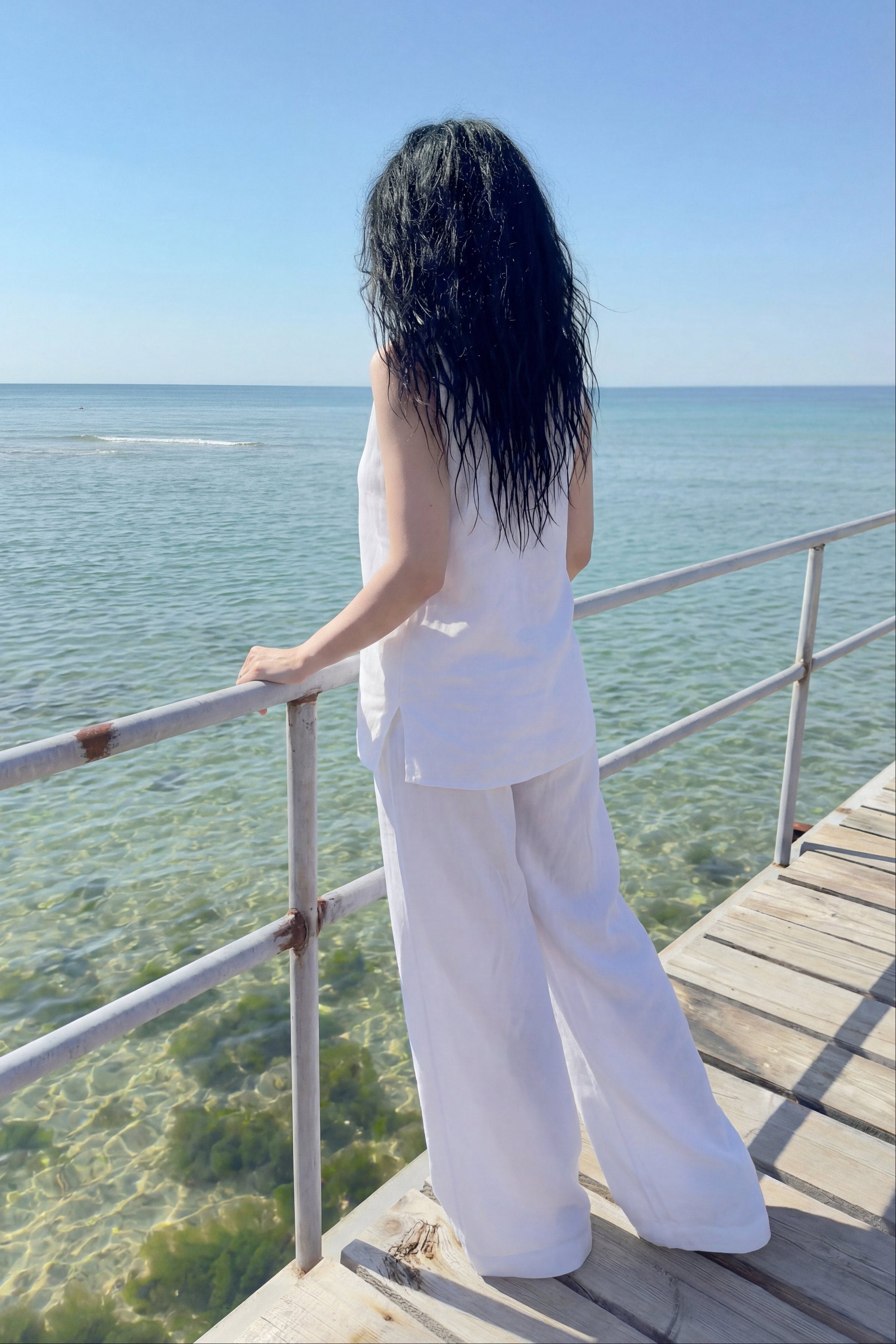 Person in white outfit standing on a wooden deck overlooking clear blue water.