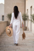 Woman in a white outfit walking with a straw hat and broom in an outdoor setting.