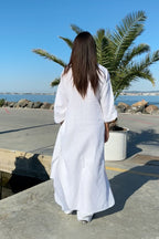 Person in a white dress standing by a waterfront with palm trees and rocks.