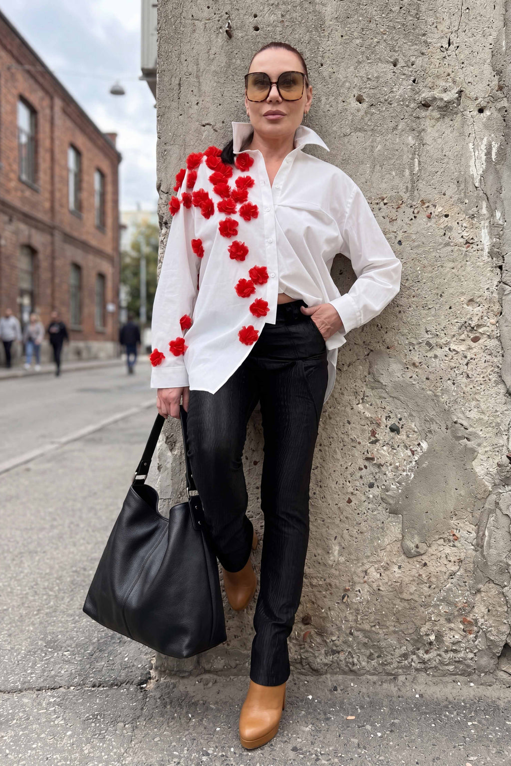 Woman in a white shirt with red floral patterns, black pants, and brown shoes standing against a textured wall.