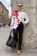 Woman in a white shirt with red floral patterns, black pants, and brown shoes standing against a textured wall.