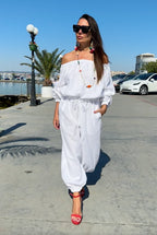 Woman in a white outfit walking on a sidewalk by a waterfront with cars and palm trees in the background.