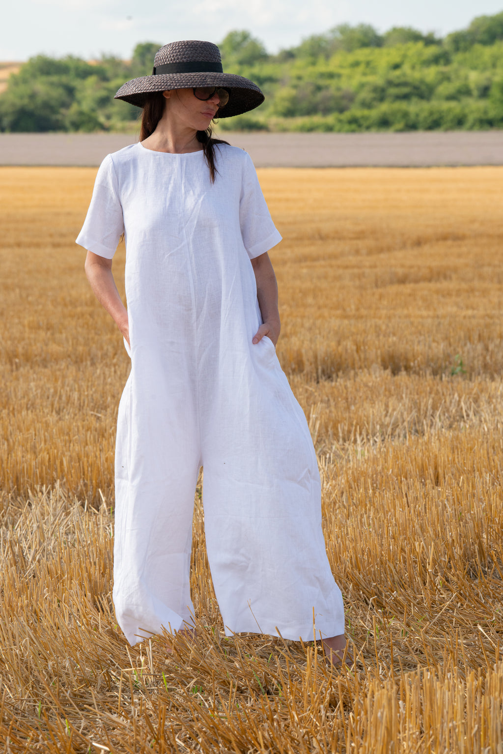 Woman in a white jumpsuit and wide-brimmed hat standing in a golden field.