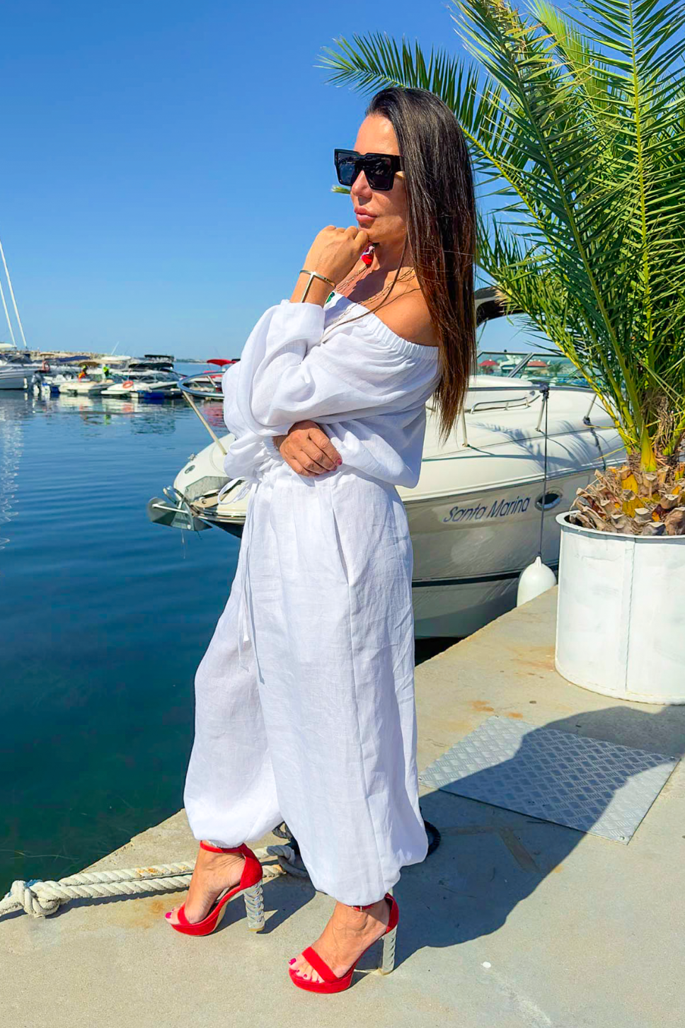 Woman in a white outfit with red sandals standing by a marina with boats and palm trees.