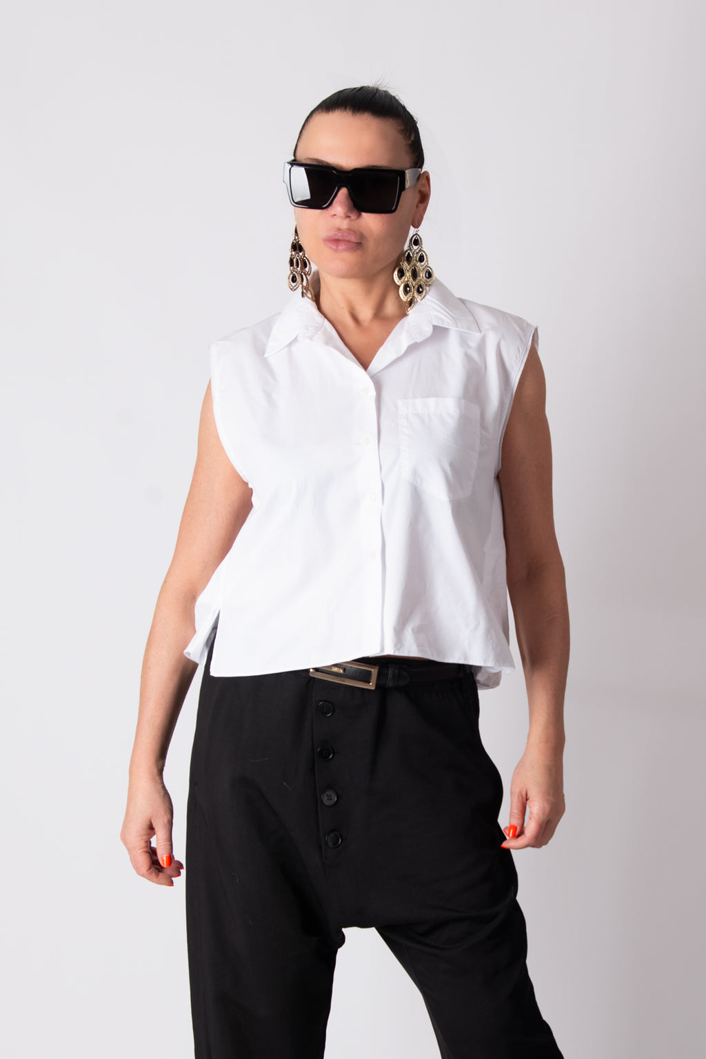 Person wearing a white sleeveless shirt and black pants with sunglasses on a plain background