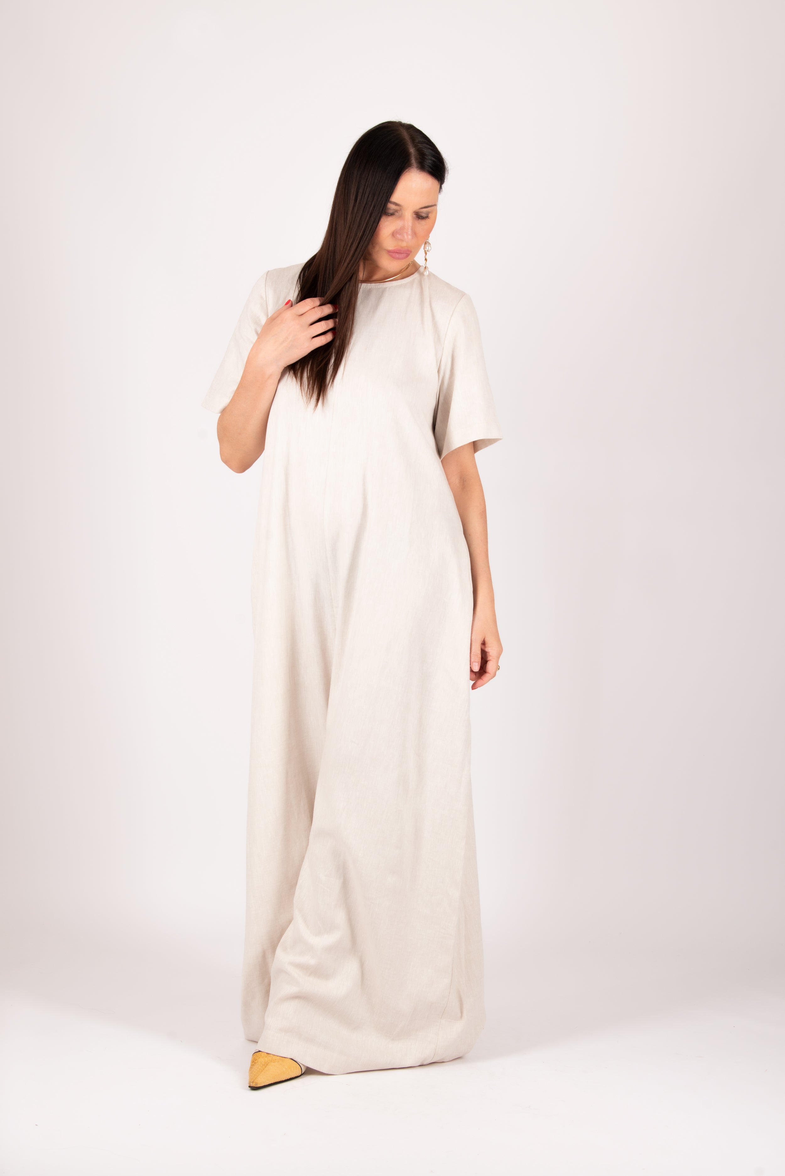 A woman modeling a loose, high-neck, short-sleeved maxi dress in a light cream or off-white linen-like fabric, standing against a plain light gray studio background.