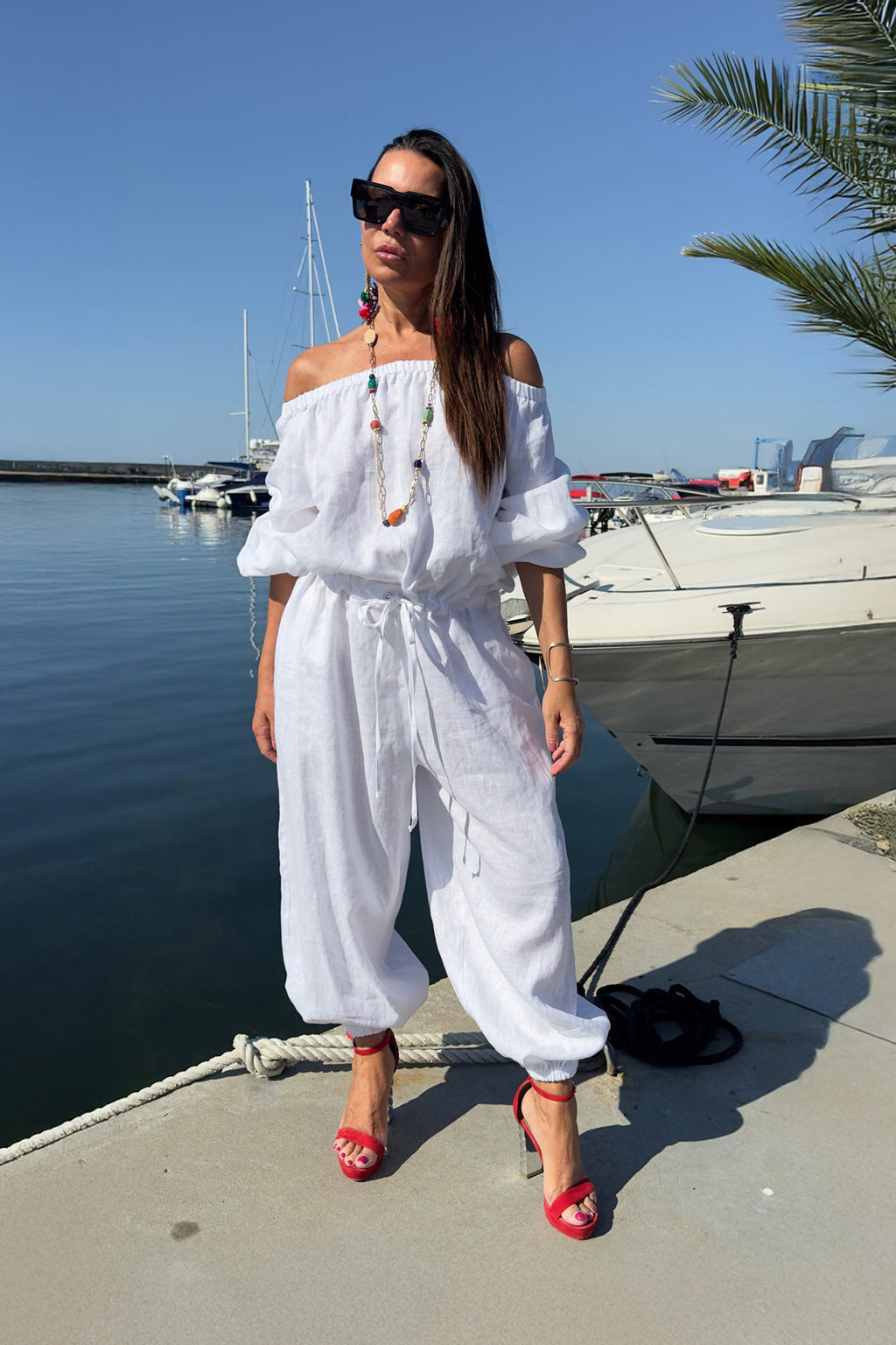 Woman in a white outfit standing by a waterfront with boats and palm trees.