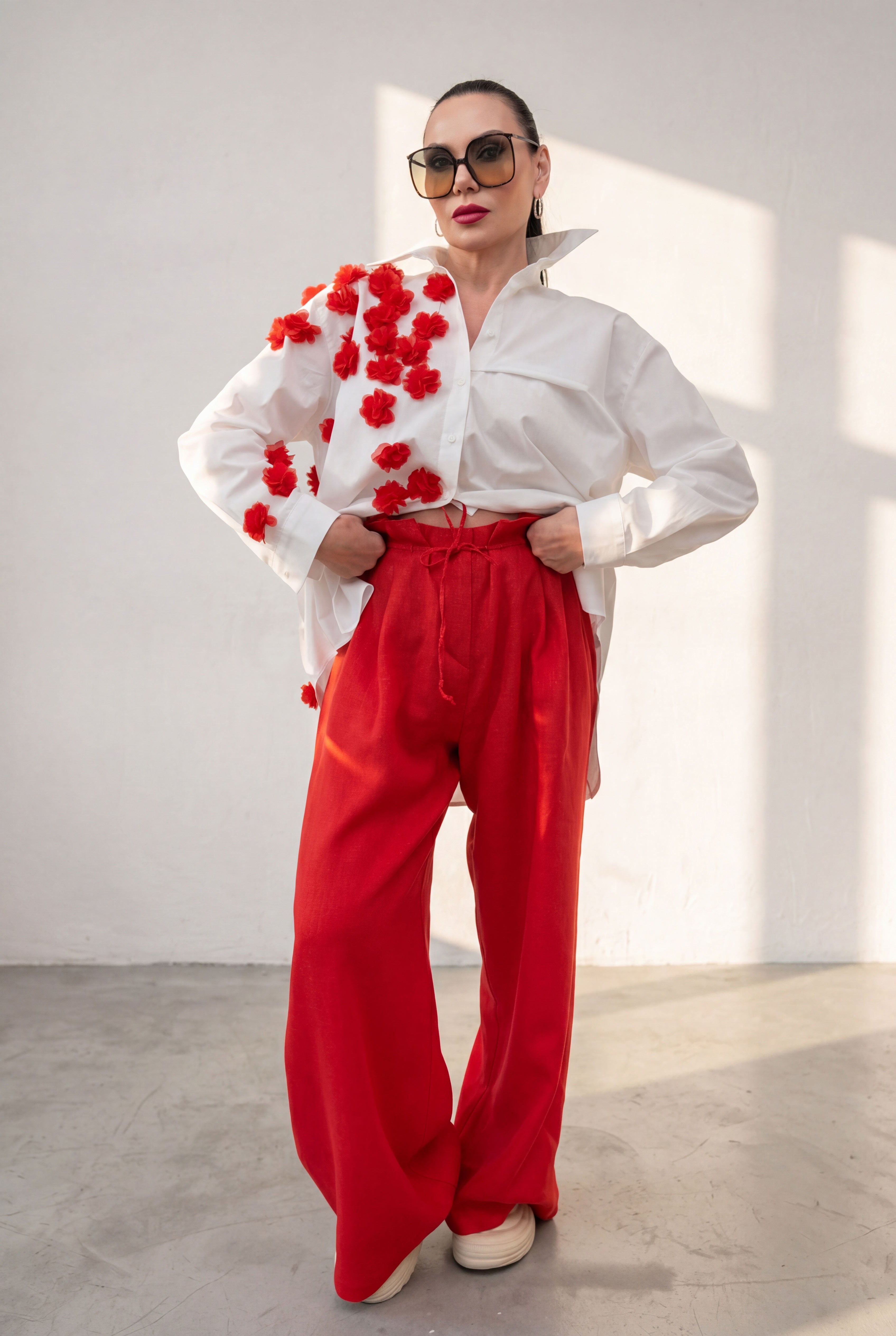 Woman wearing a white blouse with red floral embellishments and red pants against a white wall.