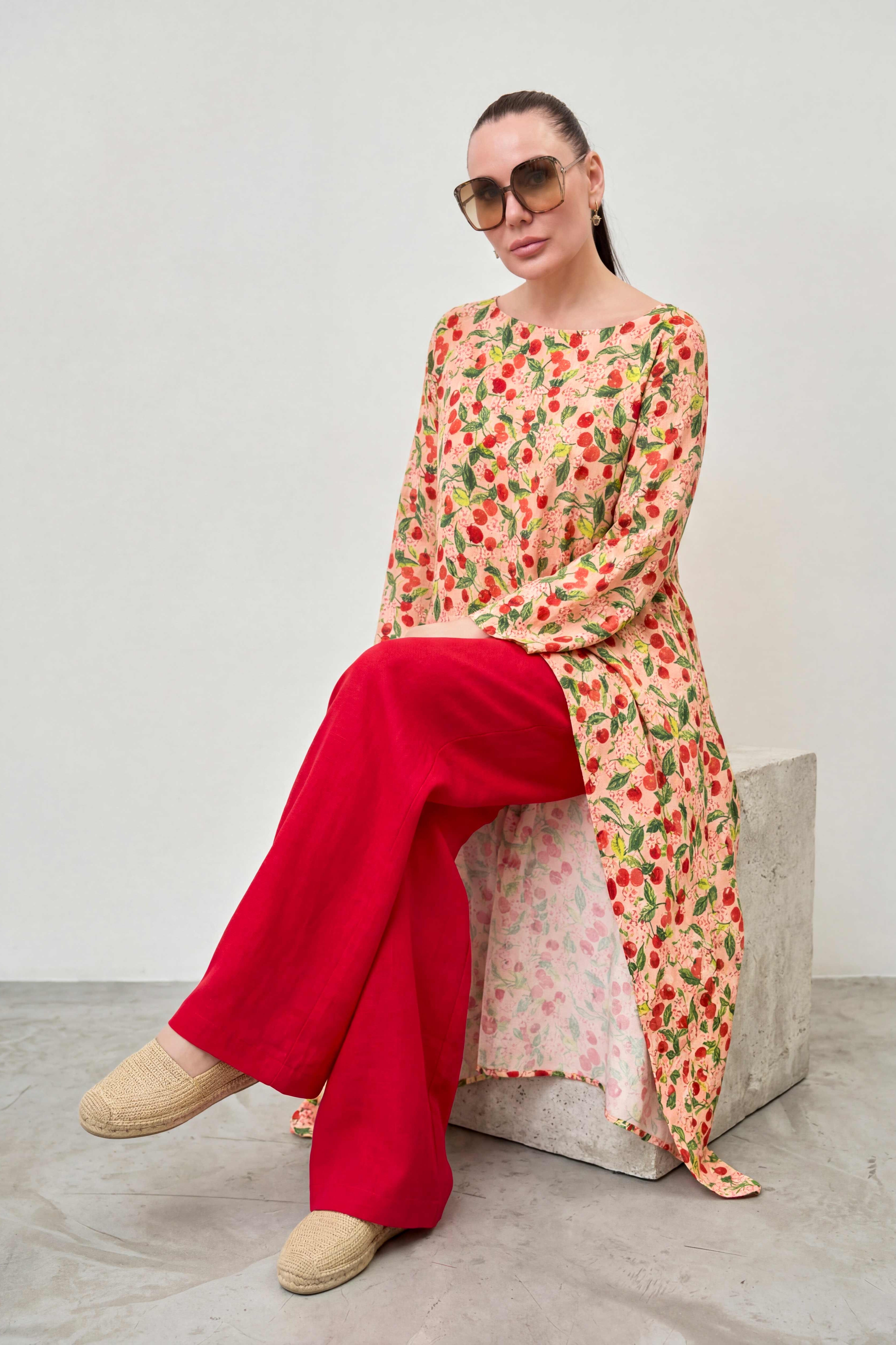 Woman wearing a floral dress with red pants sitting on a concrete block against a white wall.