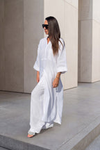 A woman wearing a flowing white maxi dress and black sunglasses walks barefoot on concrete steps against a minimalist beige stone wall.