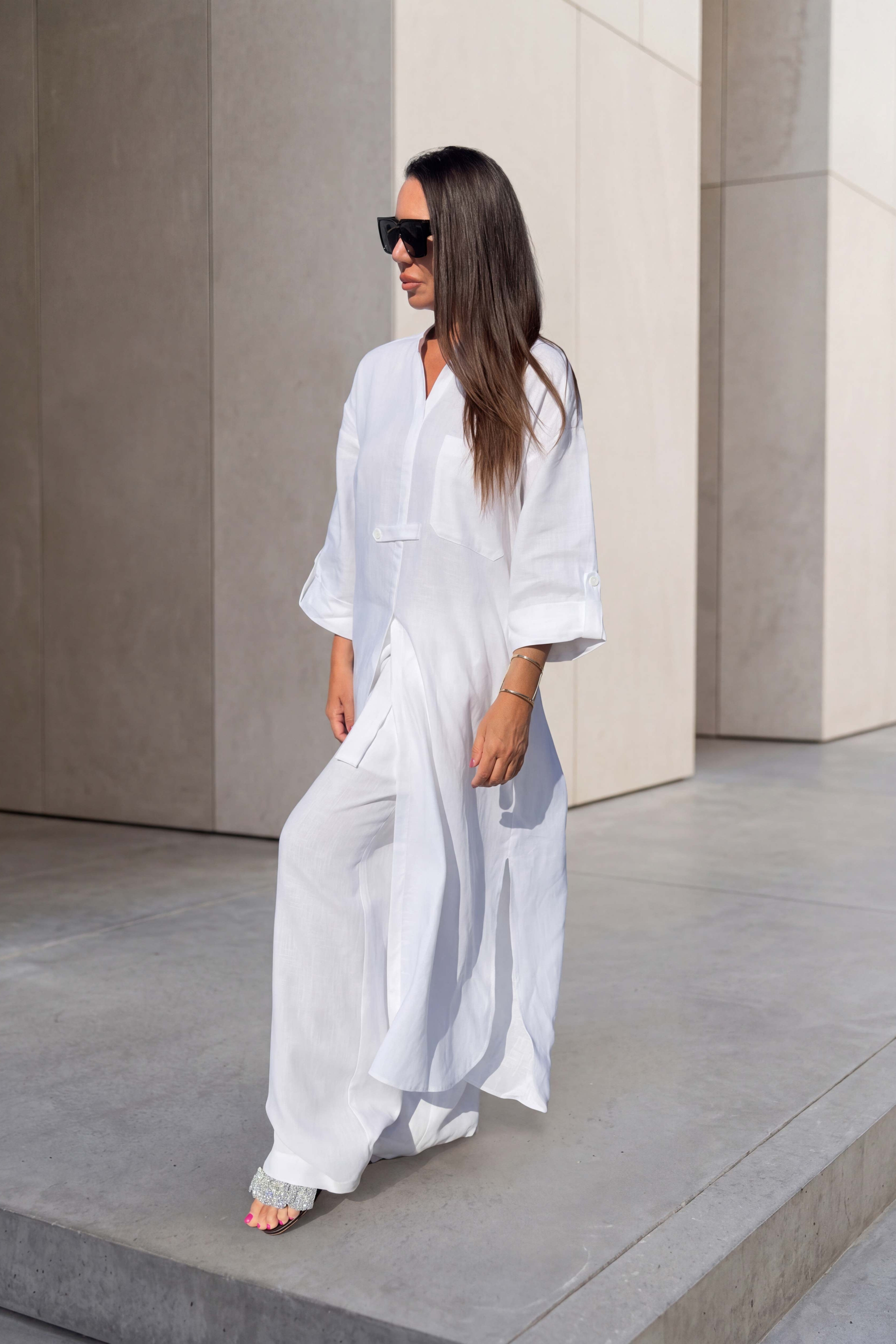 A woman wearing a flowing white maxi dress and black sunglasses walks barefoot on concrete steps against a minimalist beige stone wall.
