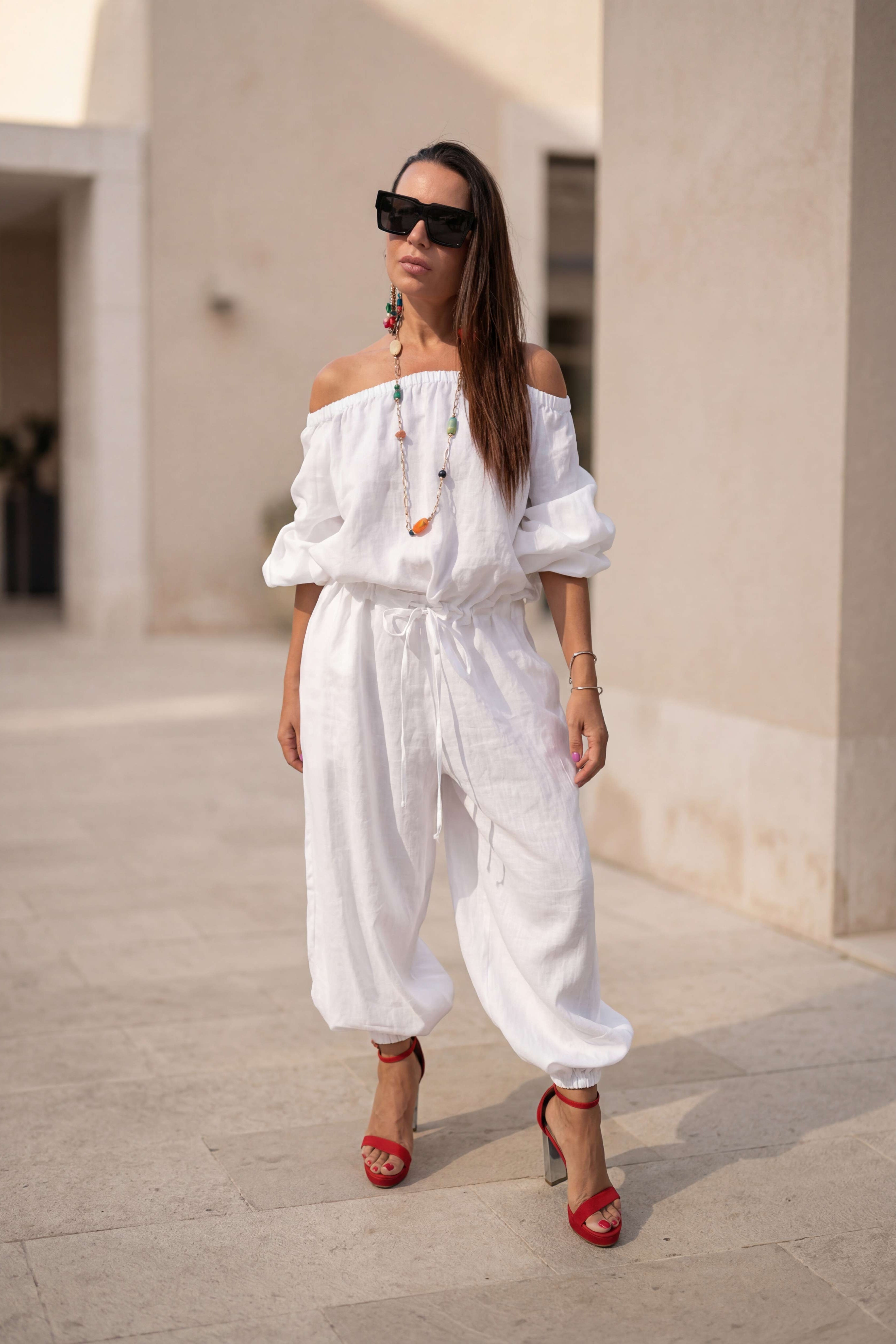 Woman in a white jumpsuit with red shoes standing on a stone pavement.