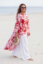 Woman in a red floral dress and white pants standing on a sandy beach.