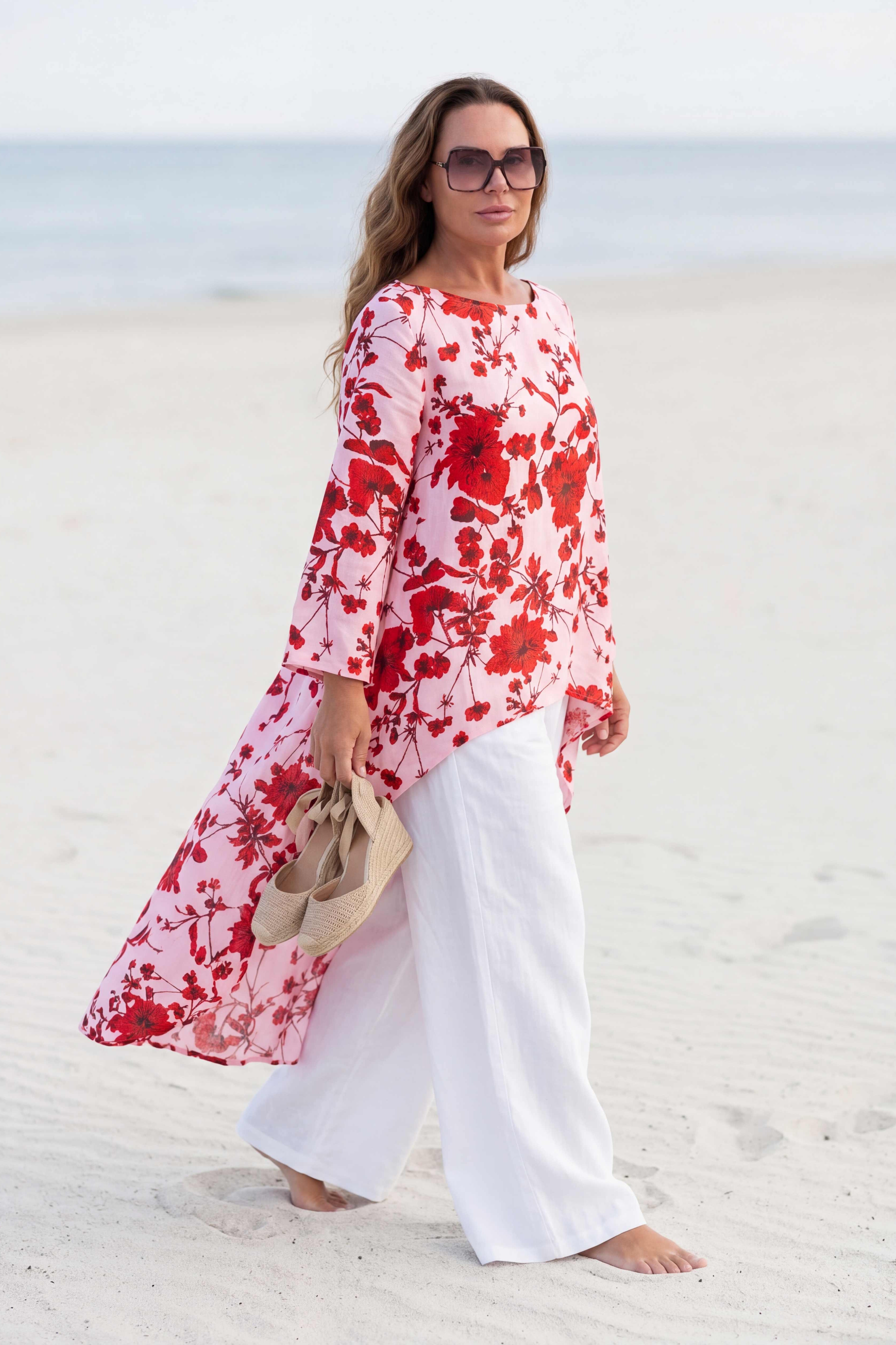 Woman in a red floral dress and white pants standing on a sandy beach.