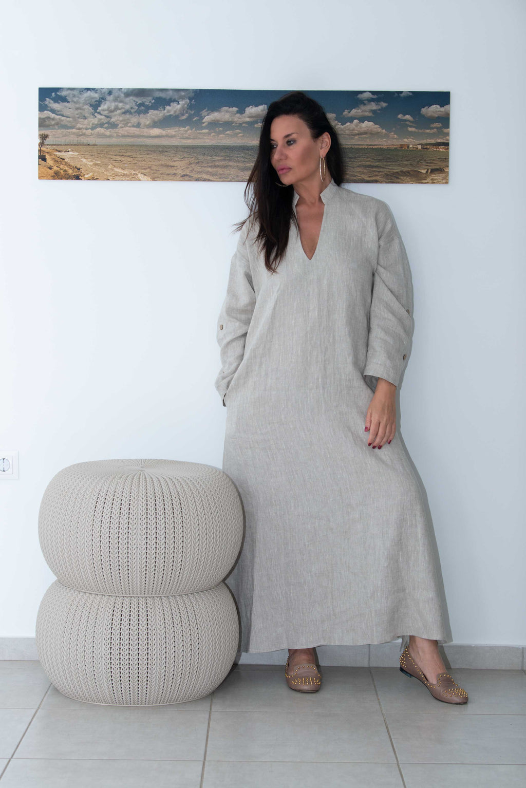 Woman in a long beige dress standing next to a textured ottoman against a white wall with a painting of a beach scene.