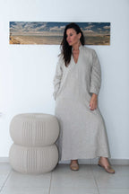 Woman in a long beige dress standing next to a textured ottoman against a white wall with a painting of a beach scene.